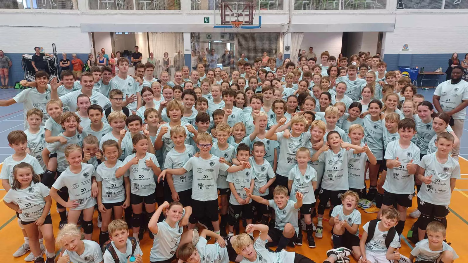Een grote groep kinderen in witte stage-shirts poseert samen in de sporthal tijdens de zomerstage