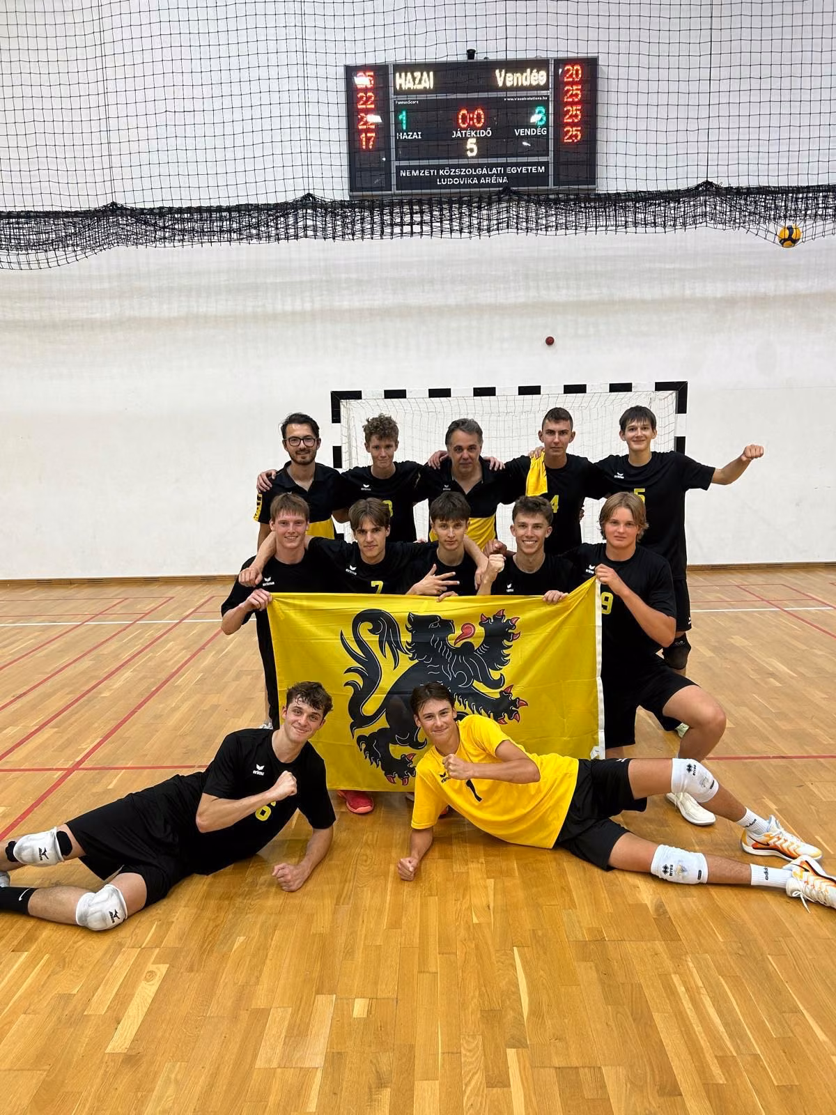 Het Vlaamse team poseert in de sporthal in Boedapest met een Vlaamse leeuwenvlag onder het scorebord.