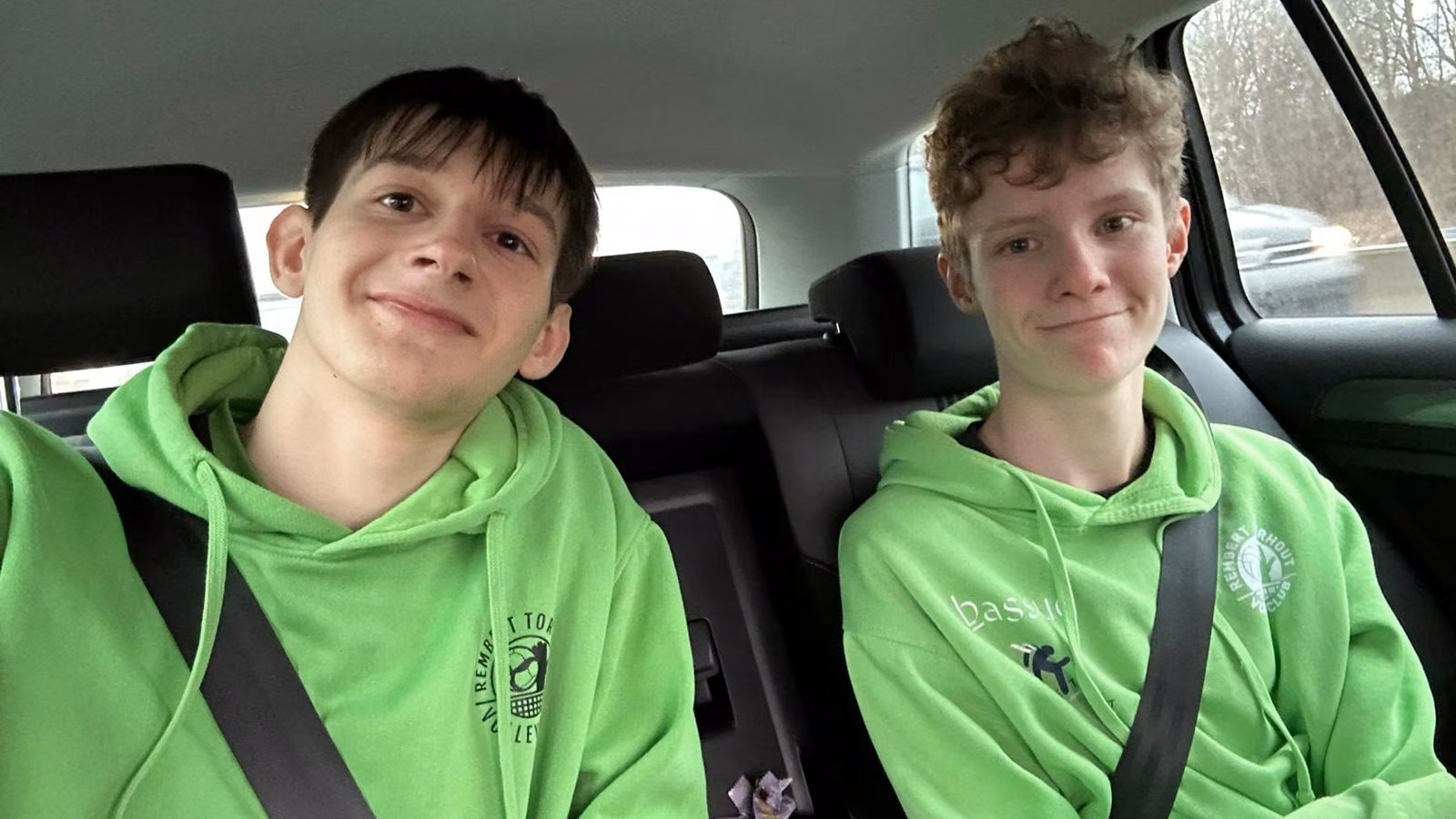 Twee jeugdspelers in een groene clubhoodie zitten glimlachend naast elkaar op de achterbank van een auto.